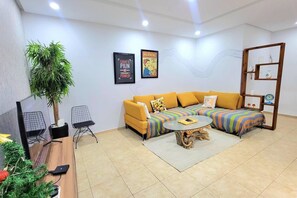 Ein modernes Wohnzimmer mit einem gelben Sofa, einem runden Couchtisch, einer großen Topfpflanze und zwei gerahmten Postern an der Wand.