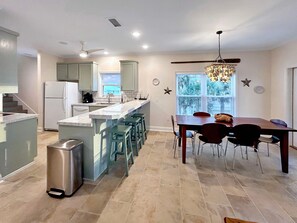 A modern kitchen with a central island, a dining area with a wooden table and chairs, and a large window with blinds. - Summer Salt (Port St. Joe)