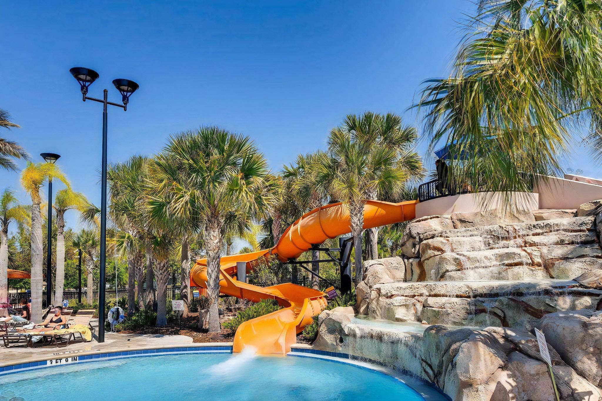 A pool area with a water slide, palm trees, and a rocky backdrop.
