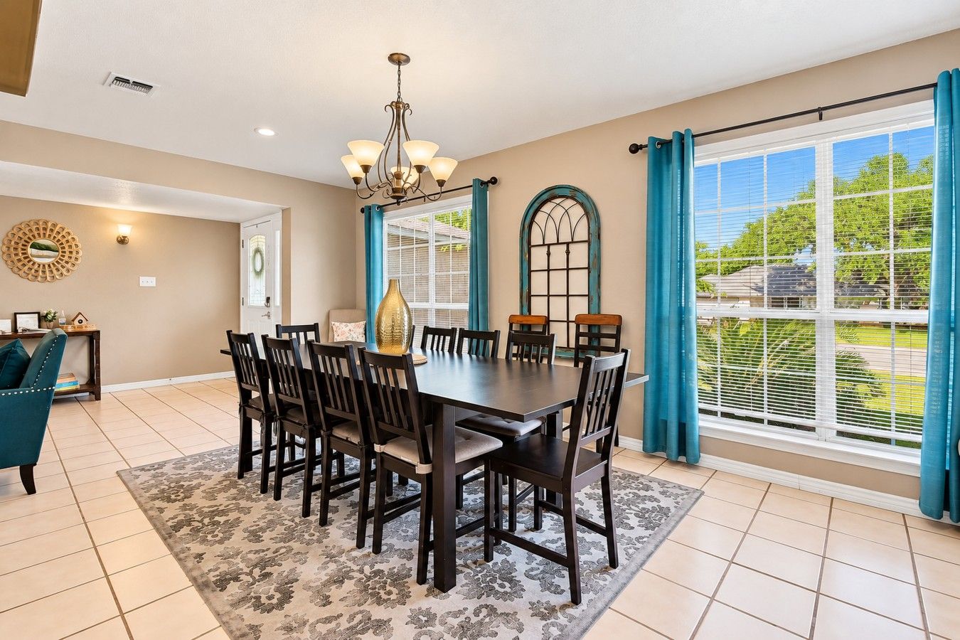 A dining room with a large table, chairs, a chandelier, and a view through the windows.