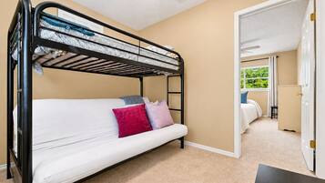 Cama litera con colchón, almohadas y cabecera. Se ve una puerta que da a otra habitación con una cama y una ventana.