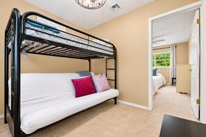 Uma cama beliche com colchão, travesseiros e cabeceira. Uma porta dá acesso a outro quarto com cama e janela.