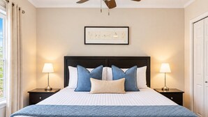 Un lit bien fait, recouvert d’une couette blanche matelassée, d’un plaid bleu matelassé et de plusieurs coussins dans différentes nuances de bleu et de blanc. Deux lampes de chevet sont placées de chaque côté du lit, et une œuvre d’art encadrée est accrochée au-dessus.
