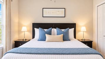 A neatly made bed with a quilted white duvet, a blue quilted throw, and multiple pillows in shades of blue and white. There are two bedside lamps on either side of the bed, and a framed artwork above the bed.