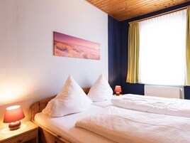Ein Schlafzimmer mit einem Bett, Kissen, einem Nachttisch mit Lampe, einem Bild an der Wand und einem Fenster mit Vorhängen.