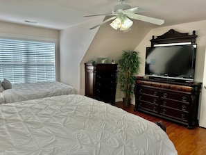 Un dormitorio con dos camas, ventilador de techo, televisor, cómoda y planta en maceta.