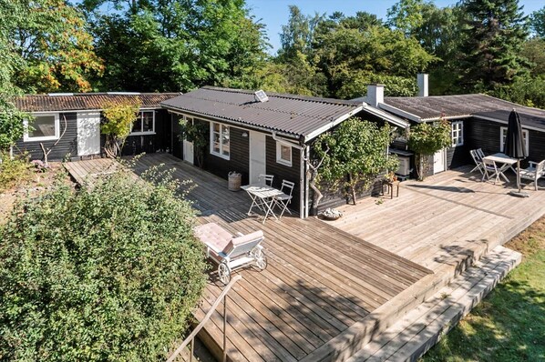 Eine Holzterrasse mit Gartenmöbeln, eine überdachte Veranda und ein Haus mit dunklem Dach und weißen Verzierungen.