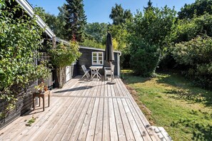 Eine Holzterrasse mit Tisch und Stühlen, umgeben von Pflanzen und einem kleinen Gebäude.