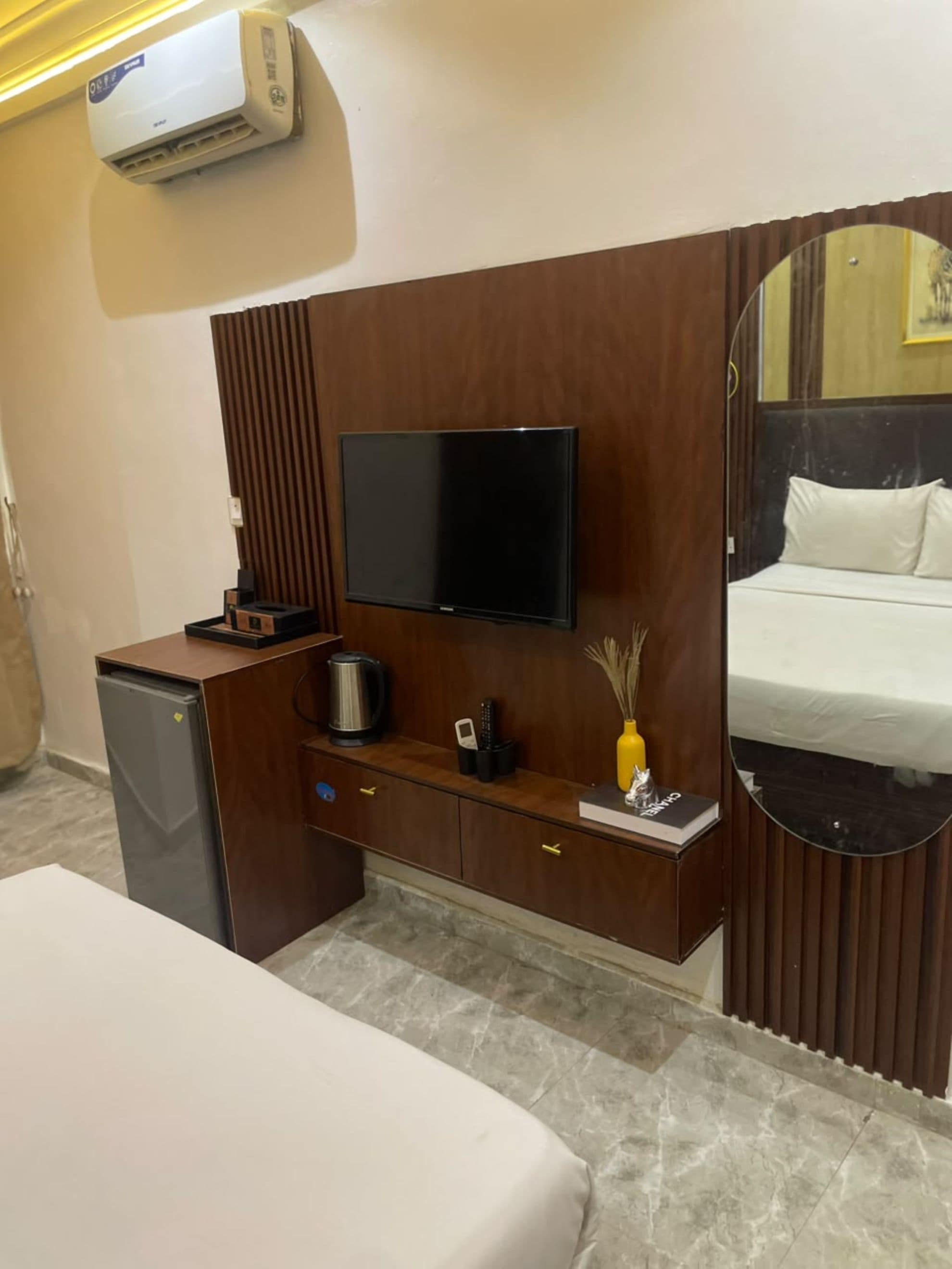 A hotel room with a bed, a TV mounted on a wooden panel, a small refrigerator, a coffee maker, and a bedside table with a vase.