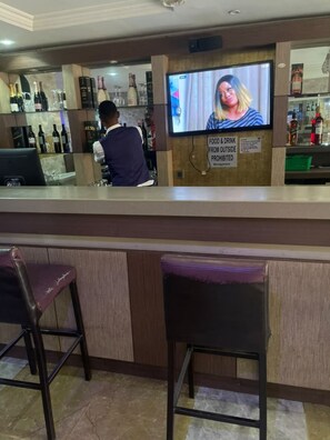 Zona de bar con una pantalla de televisión, un cartel que prohíbe alimentos y bebidas del exterior, y un cantinero.