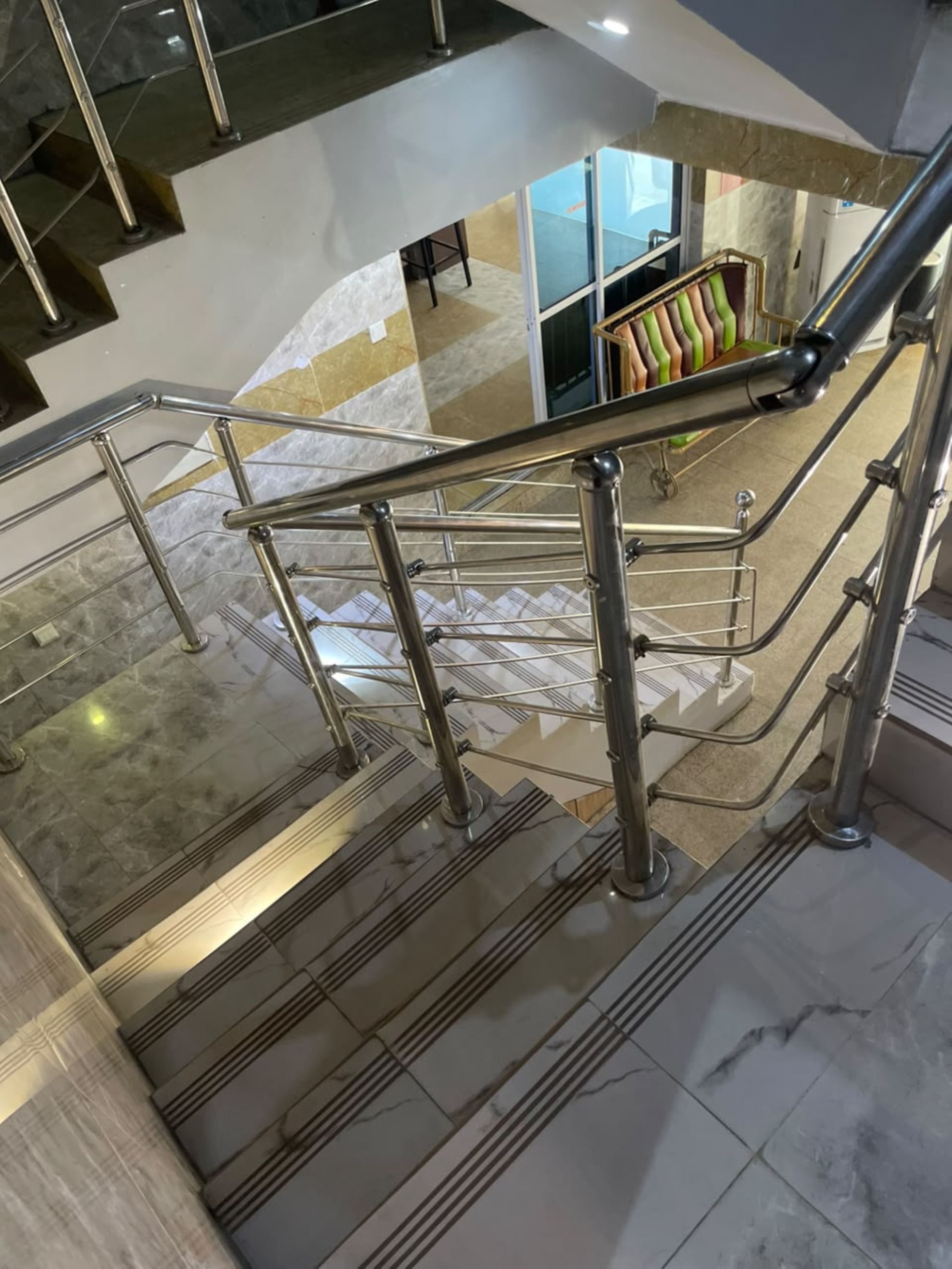 A modern interior with a glass railing, marble flooring, and a view of a seating area with colorful cushions.