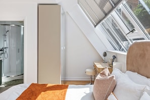 A bedroom with a skylight, a bed with pillows, a bedside table with a lamp, and a bathroom with a shower. - Stunning High-Spec 2-Bedroom Holiday Apartment in the Heart of Brighton. (Brighton and Hove)