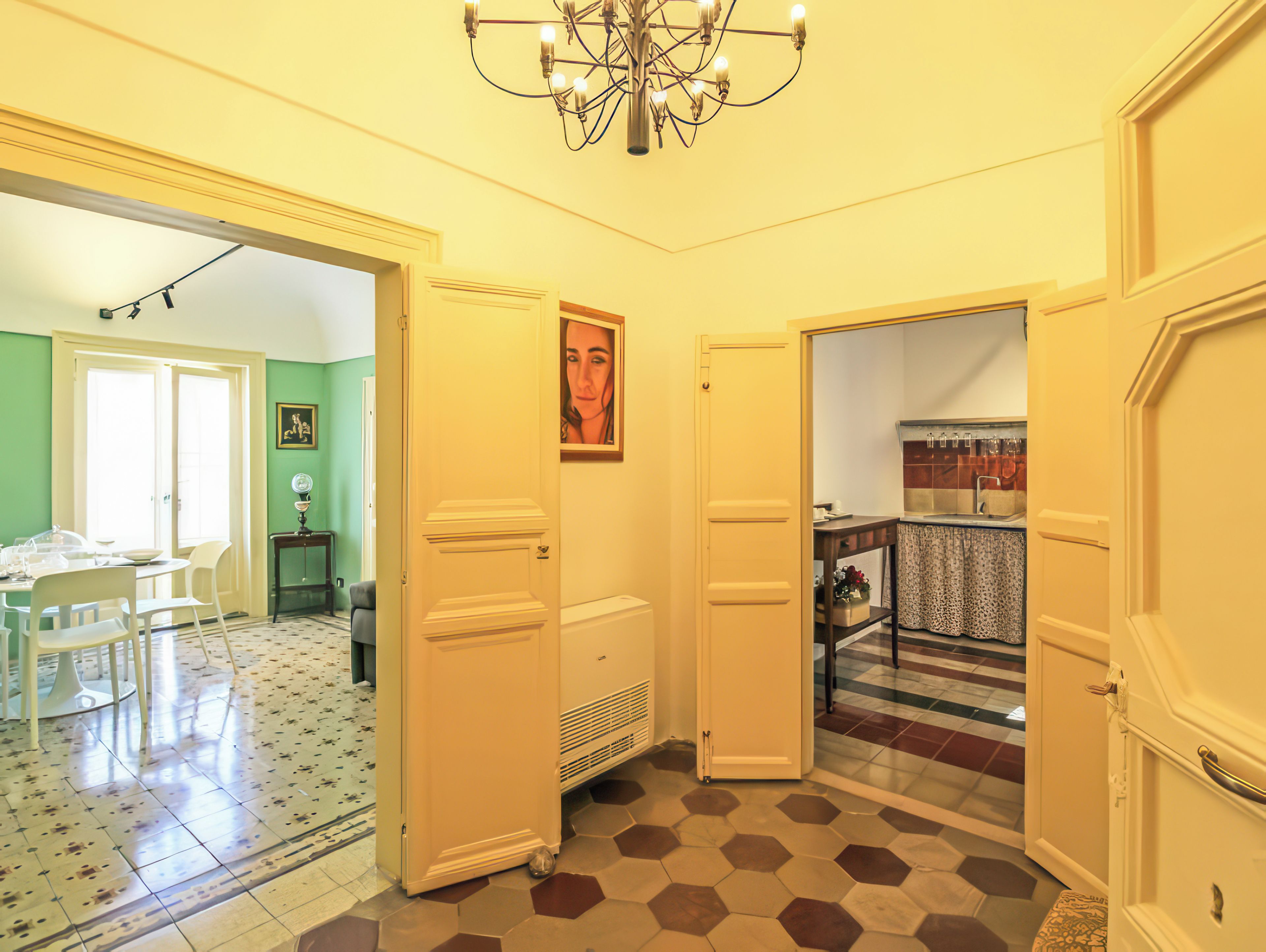 A hallway with a checkered floor, a chandelier, and a door leading to a room with a tiled backsplash.