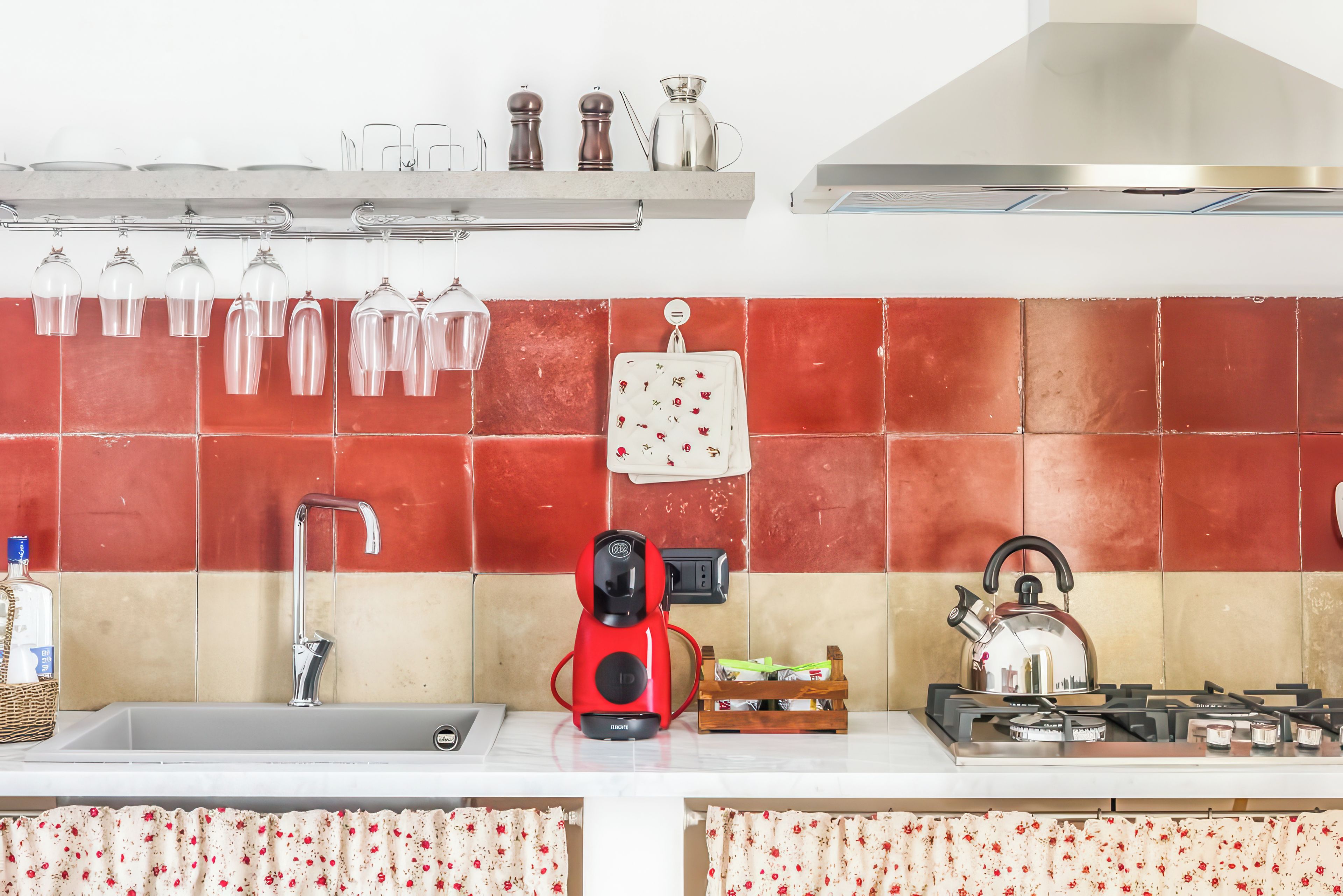 Una cocina con un salpicadero de azulejos rojos y beige, una estufa a gas, una tetera, una cafetera roja y un tendedero con vasos.
