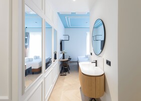 A modern bathroom with a large mirror, a round wooden vanity, and a bed in the adjacent room.