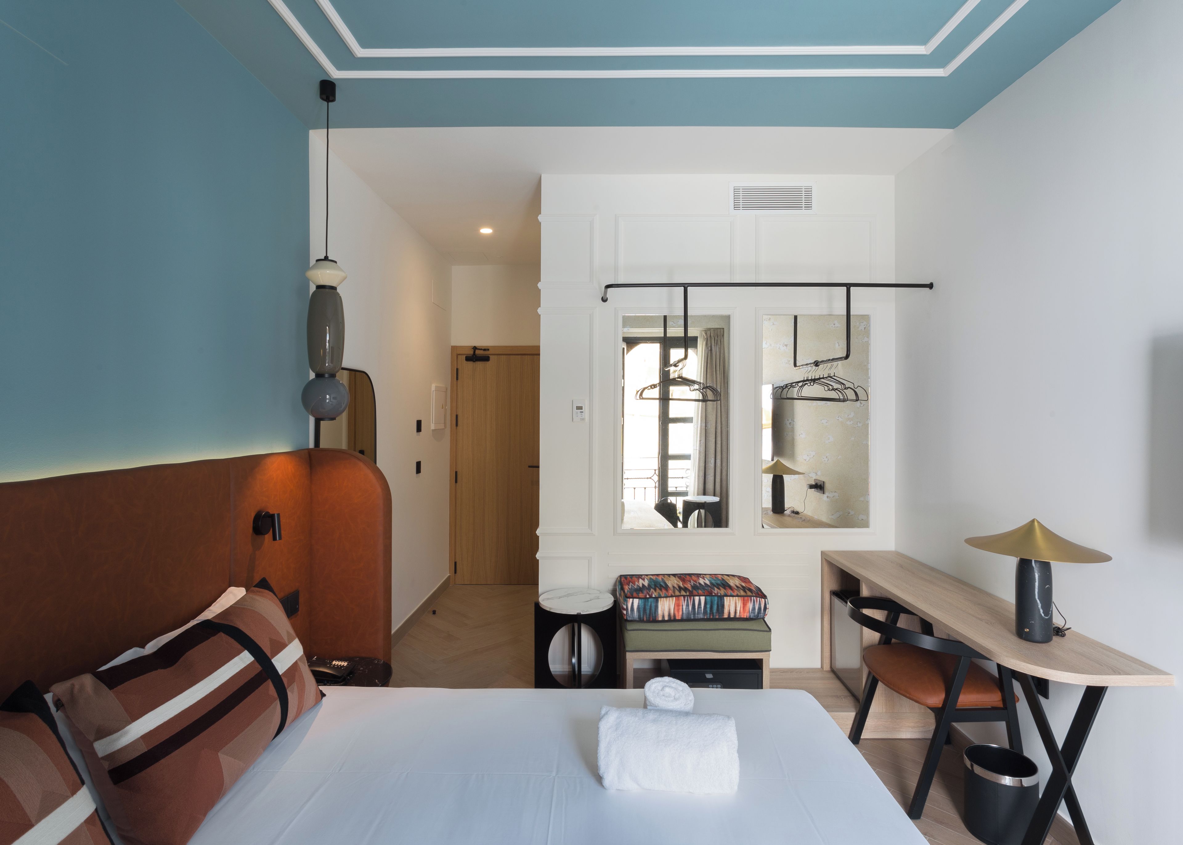 A hotel room with a bed, a desk, a chair, a bench, a hanging light fixture, and a wardrobe.