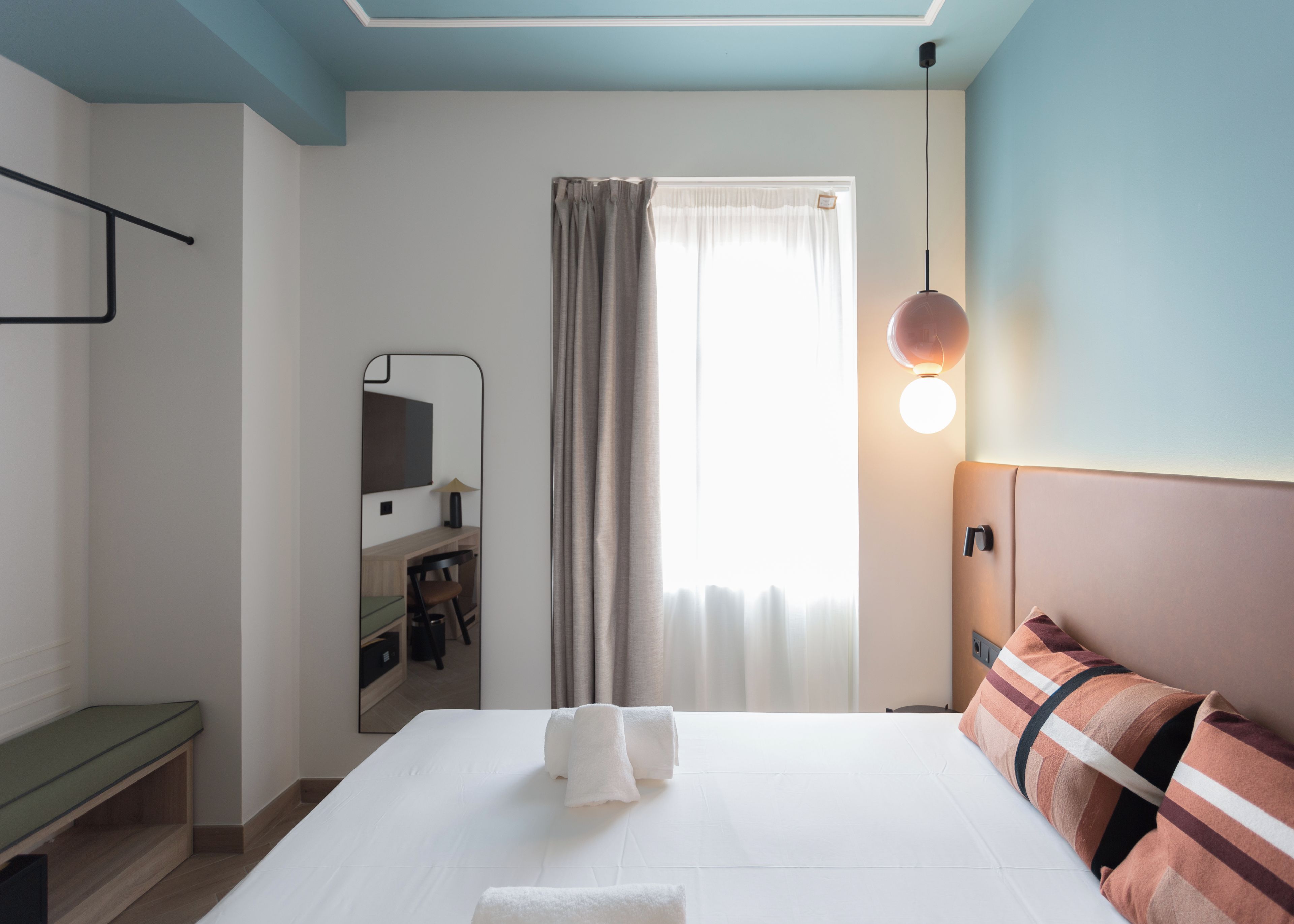 Ein Hotelzimmer mit einem Bett, einem Nachttisch, einem Spiegel, einem Schreibtisch mit Stuhl, einem Fenster mit Vorhängen und einer Wandleuchte.