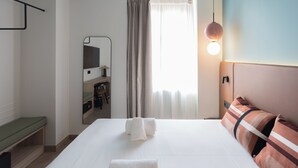 A hotel room with a bed, a bedside table, a mirror, a desk with a chair, a window with curtains, and a wall-mounted light fixture. - Casa Candela Sevilla (Sevilla)