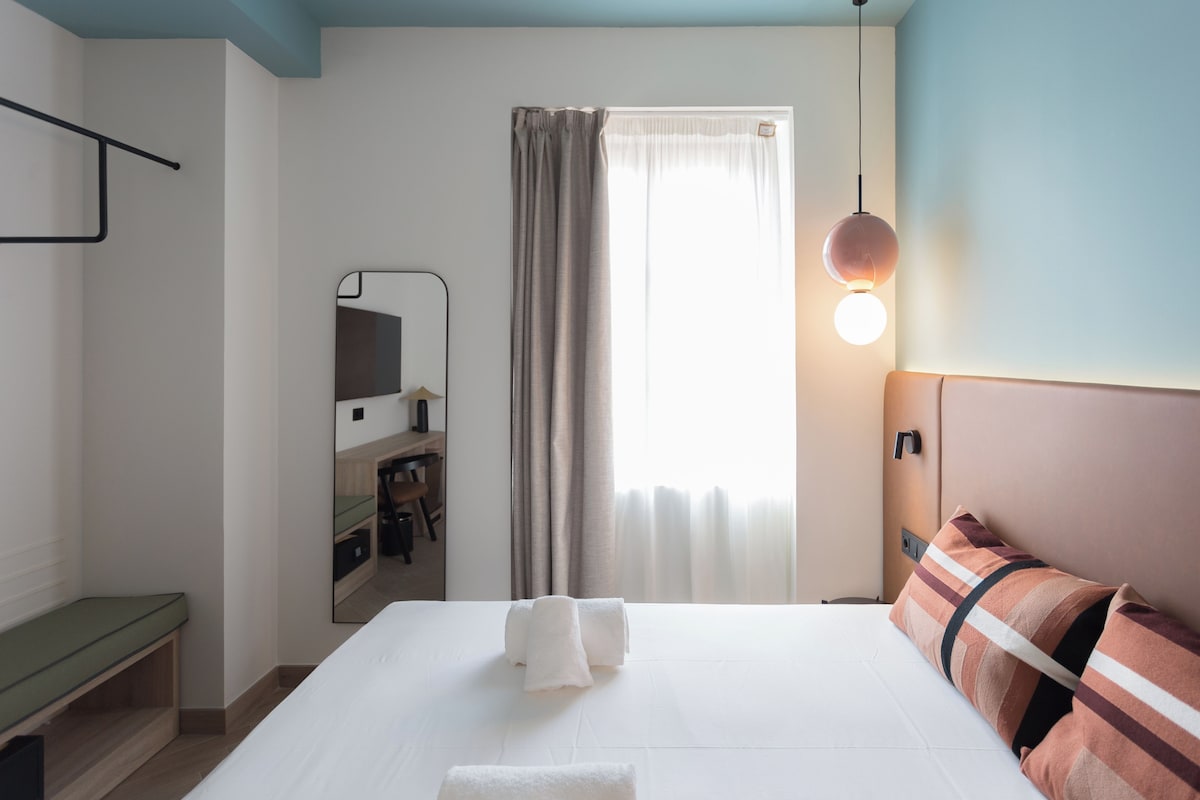 Ein Hotelzimmer mit einem Bett, einem Nachttisch, einem Spiegel, einem Schreibtisch mit Stuhl, einem Fenster mit Vorhängen und einer Wandleuchte.