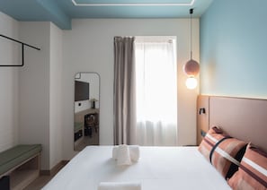 Ein Hotelzimmer mit einem Bett, einem Nachttisch, einem Spiegel, einem Schreibtisch mit Stuhl, einem Fenster mit Vorhängen und einer Wandleuchte.