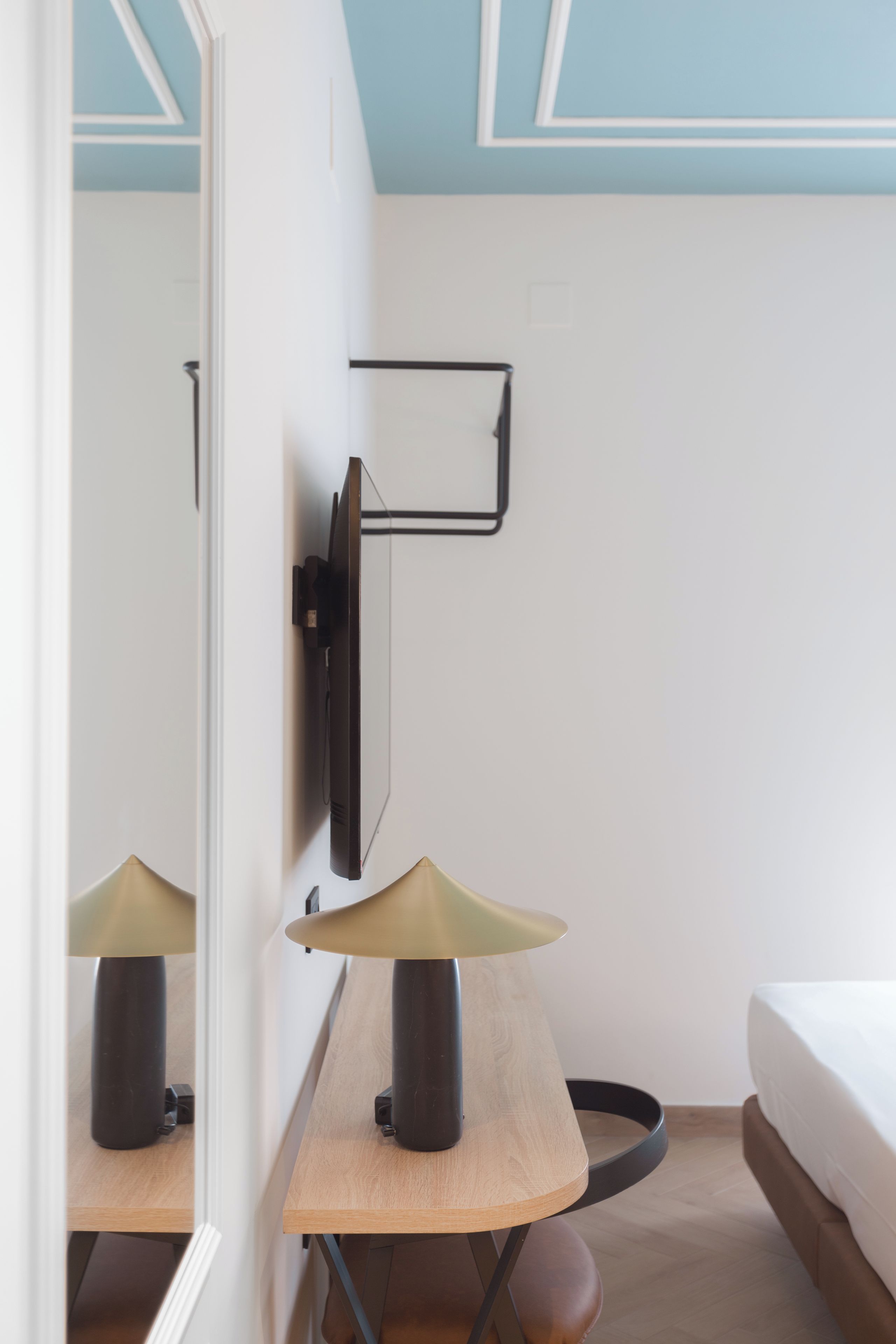 A hotel room with a bed, a desk with a lamp, a wall-mounted TV, and a mirror.