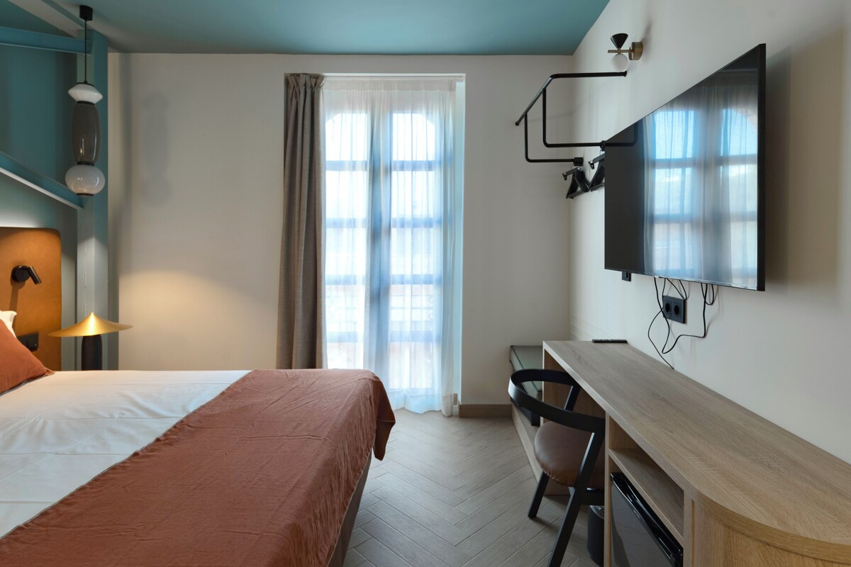 Habitación de hotel con cama, escritorio, silla, televisor y cortinas.