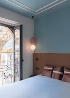 Una camera d'albergo con un letto, una lampada a sospensione e vista sui palazzi attraverso una finestra.