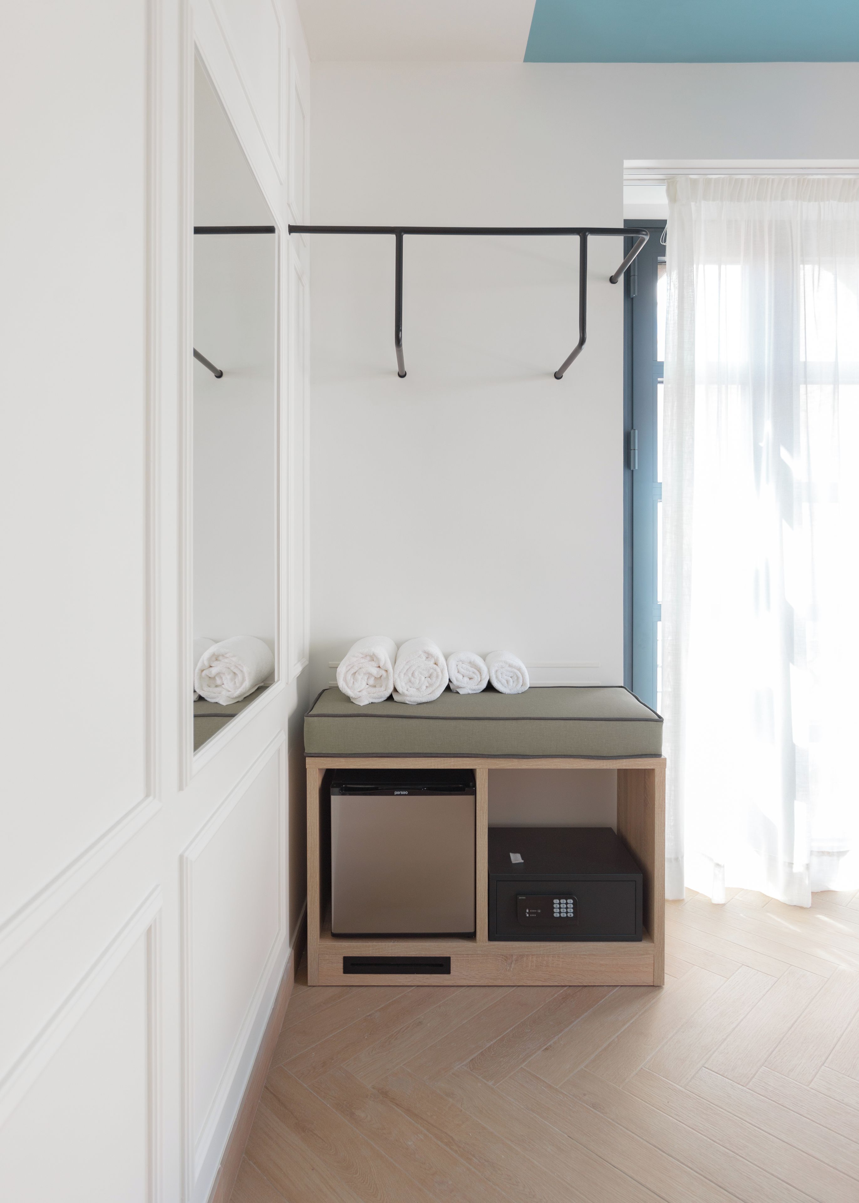 Ein Zimmer mit integriertem Kleiderschrank, einer kleinen Sitzecke, Handtüchern, einem kompakten Geschirrspüler und einem Fenster mit leichtem Vorhang.