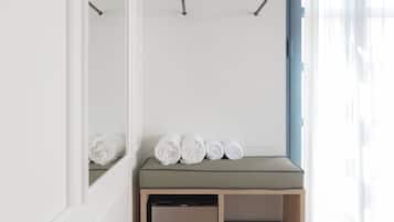 Ein Zimmer mit integriertem Kleiderschrank, einer kleinen Sitzecke, Handtüchern, einem kompakten Geschirrspüler und einem Fenster mit leichtem Vorhang.