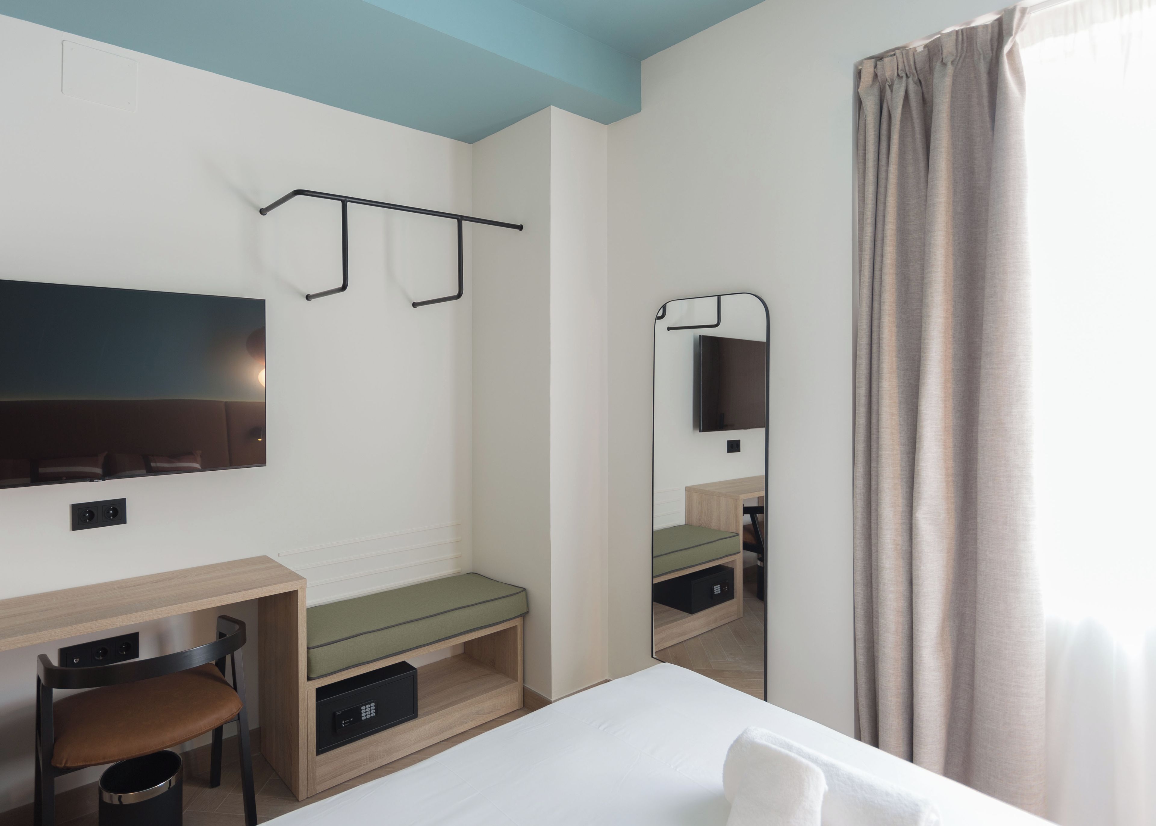 A hotel room with a bed, a desk, a chair, a TV mounted on the wall, and a mirror.