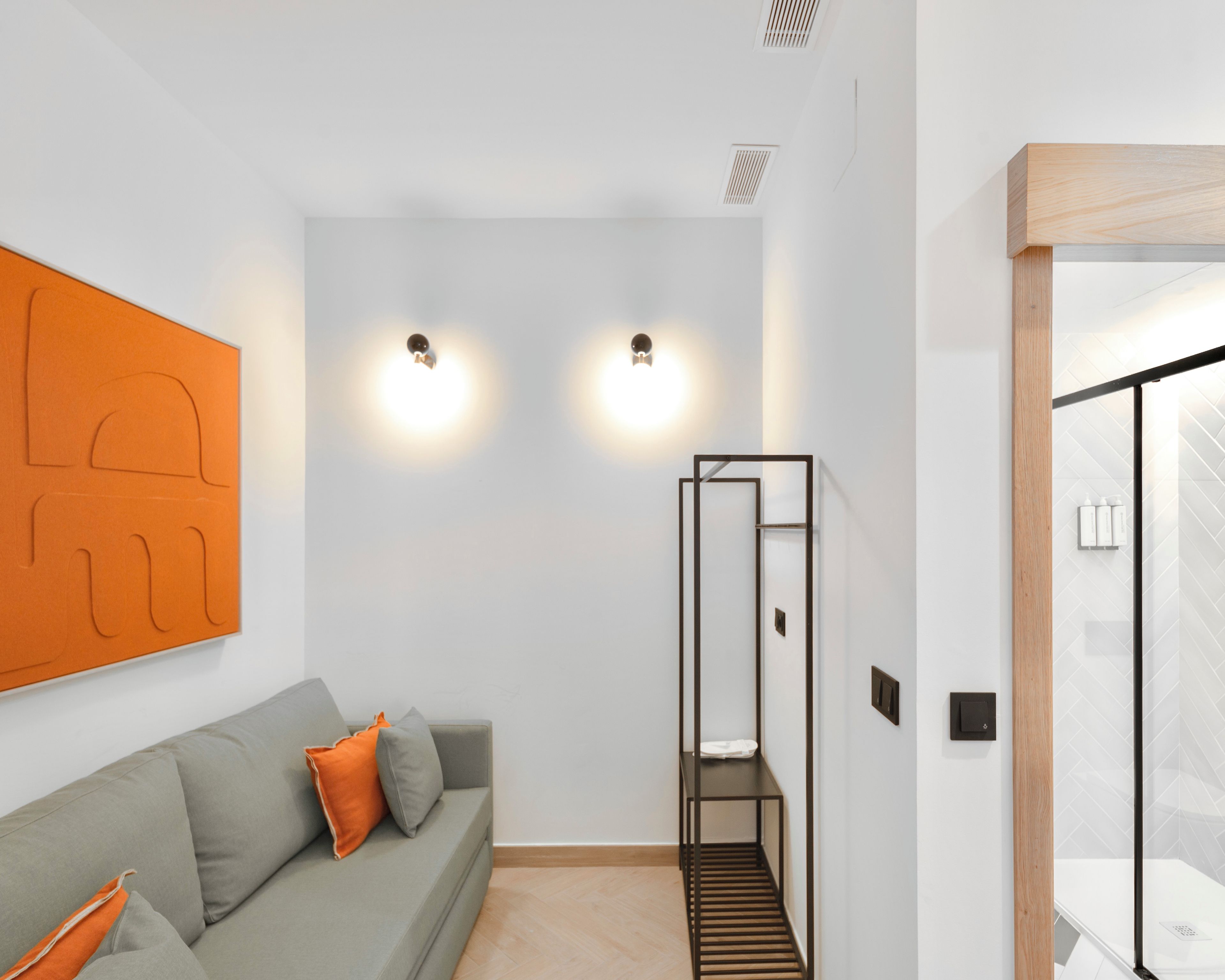 A modern hotel room with a grey sofa, two wall-mounted lights, a wooden-framed ladder, and a tiled floor.