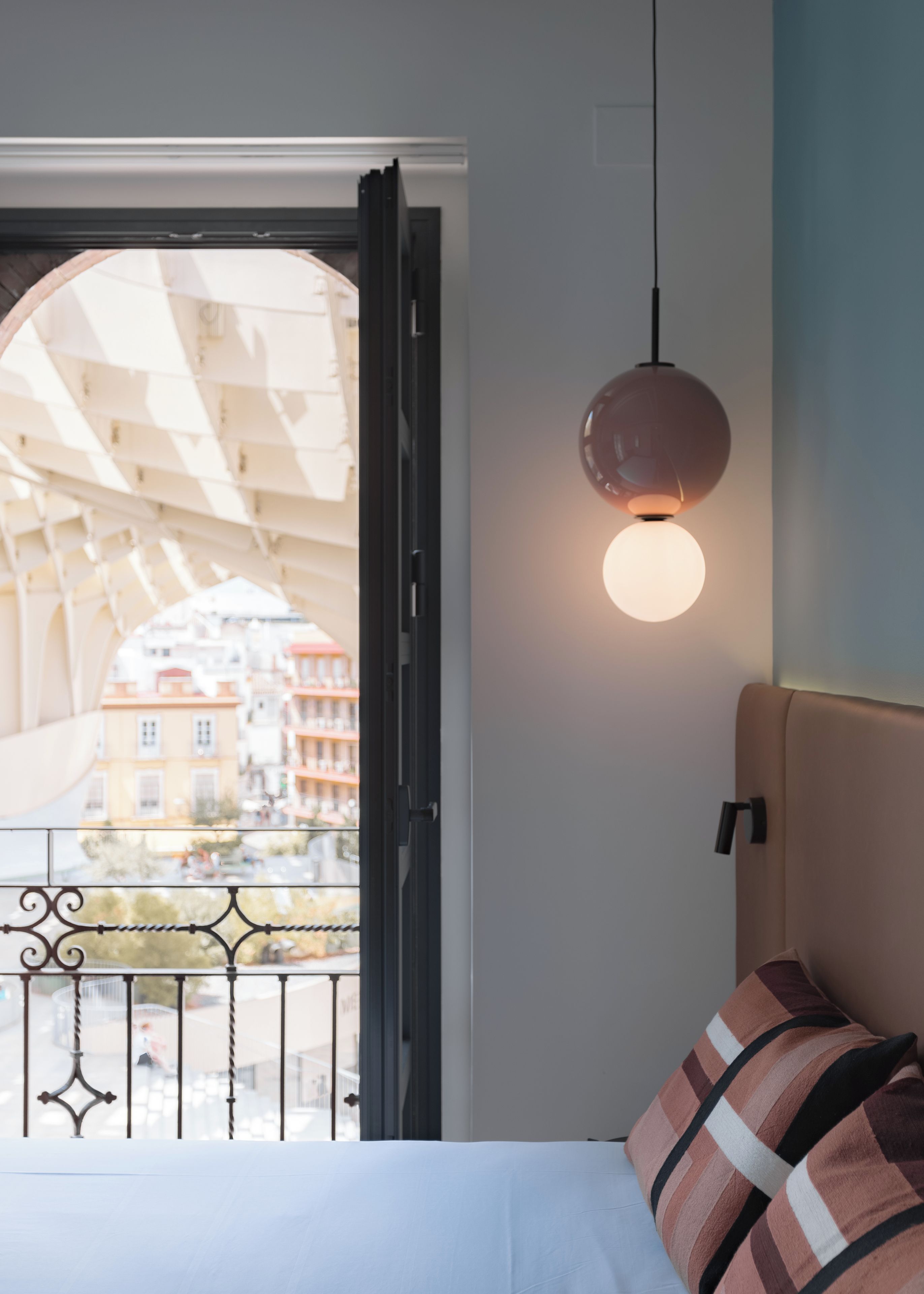 Ein Hotelzimmer mit Bett, einer Hängelampe und Blick auf Gebäude durch ein offenes Fenster.