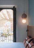Habitación de hotel con cama, lámpara colgante y vista a edificios a través de una ventana abierta.