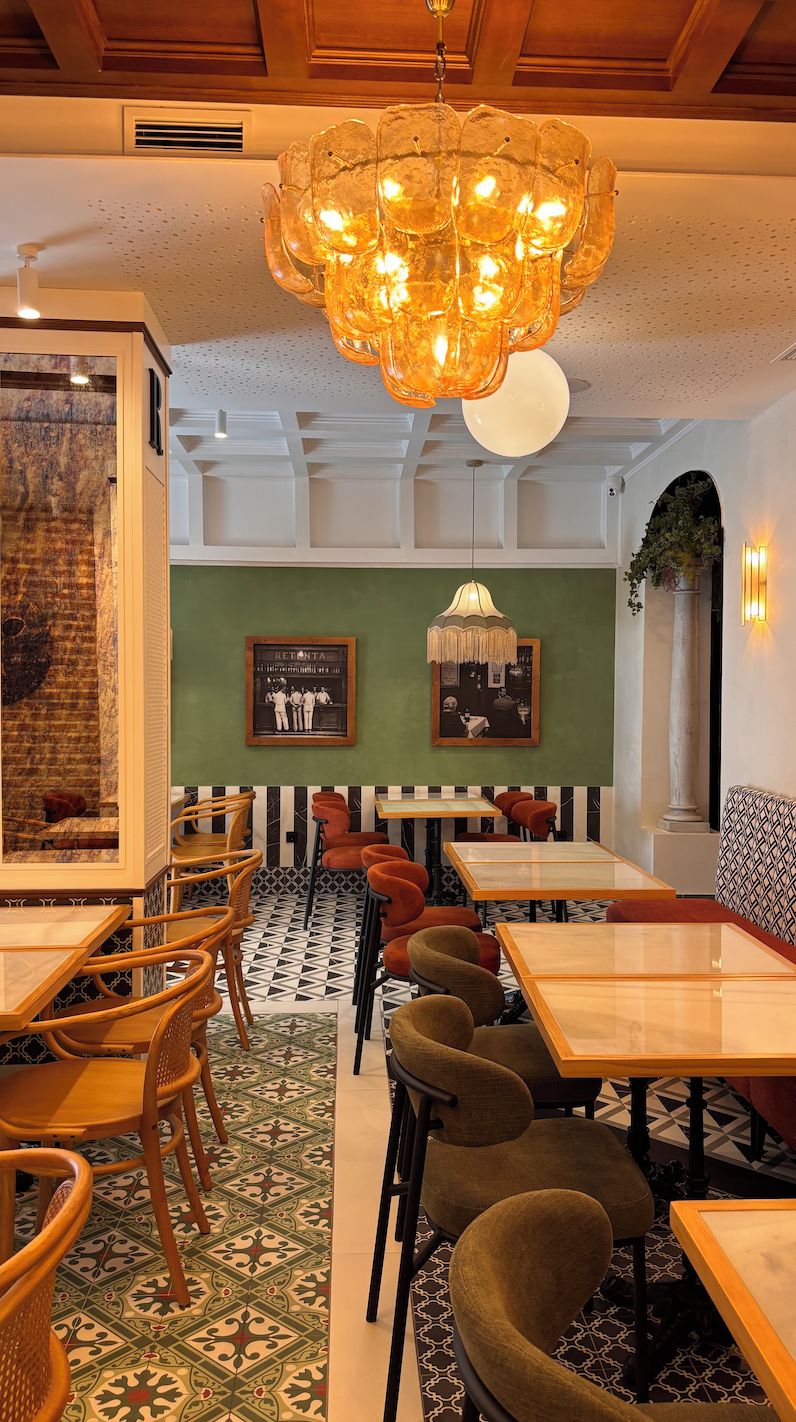 A restaurant interior with wooden tables and chairs, a patterned floor, a chandelier, and framed pictures on the wall.