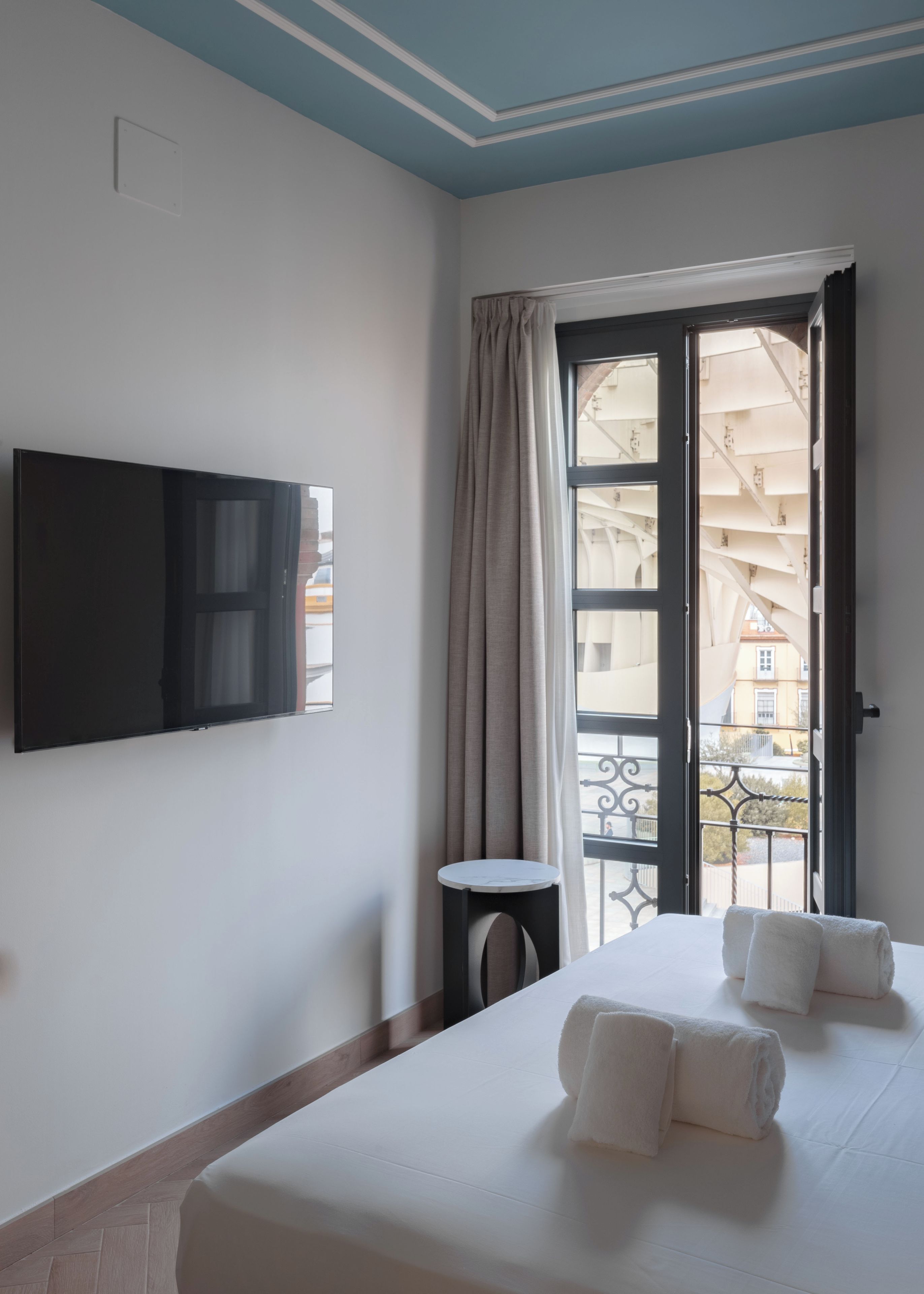 Une chambre d’hôtel avec un lit, un téléviseur fixé au mur, une petite table et un balcon donnant sur des immeubles.