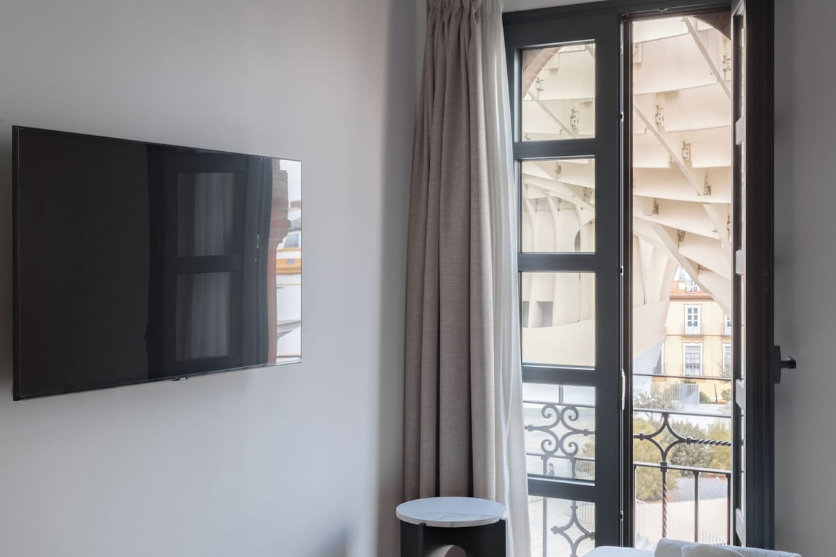 Ein Hotelzimmer mit Bett, Wandfernseher, kleinem Tisch und einem Balkon mit Blick auf Gebäude.