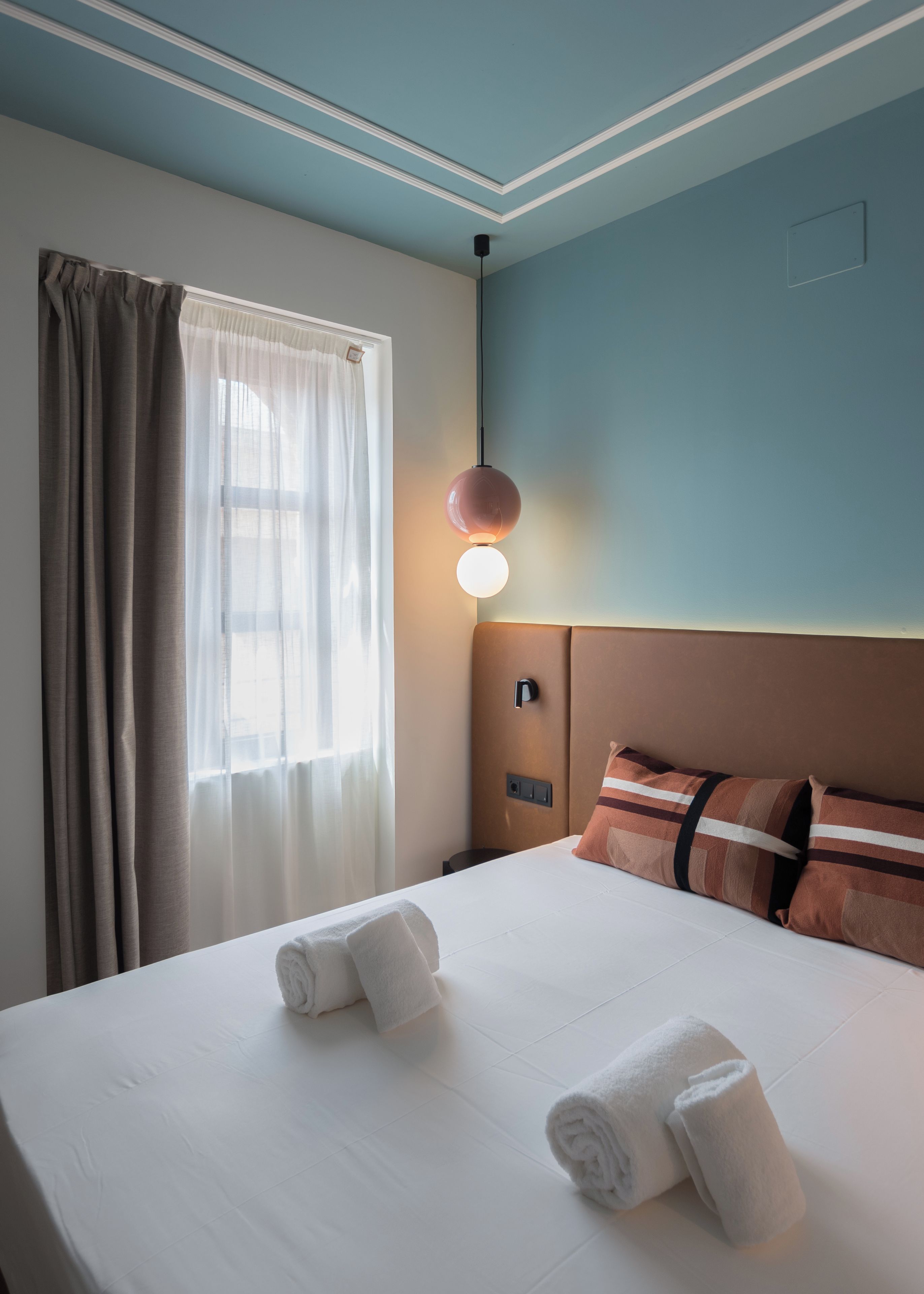Una habitación de hotel con cama, dos almohadas, mesita de noche, lámpara y ventana con cortinas.