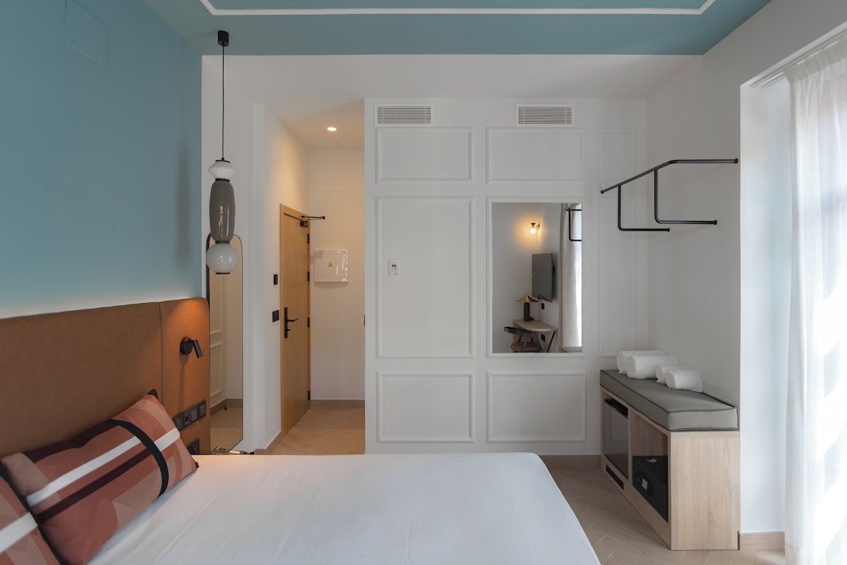 Ein Hotelzimmer mit einem Bett, einem Schreibtisch mit Spiegel, einer kleinen Küchenzeile und einem durch eine offene Tür einsehbaren Badezimmer.