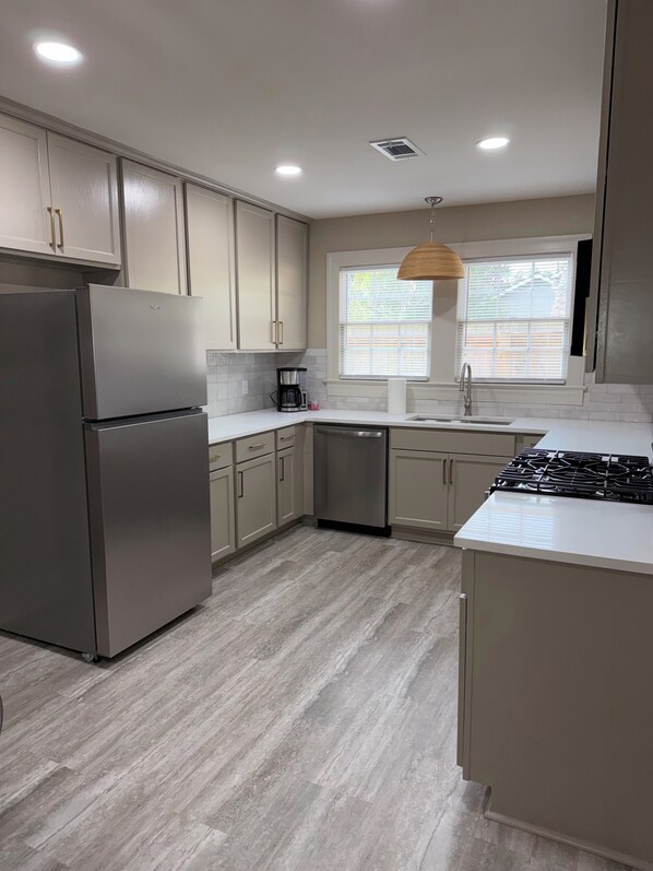 A modern kitchen with a stainless steel refrigerator, a dishwasher, a gas stove, and a window with blinds.