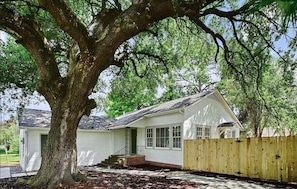 Una casa de un piso con un árbol grande en el frente, una valla de madera y un cielo despejado.