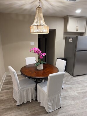 A dining area with a round table, white chairs, a hanging light fixture, and a refrigerator in the background.