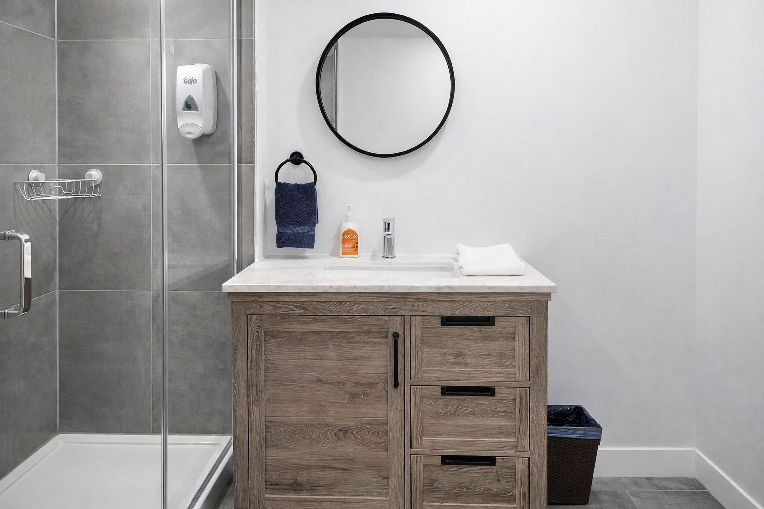 A modern bathroom with a shower, a wooden vanity, a round mirror, and a hand soap dispenser.