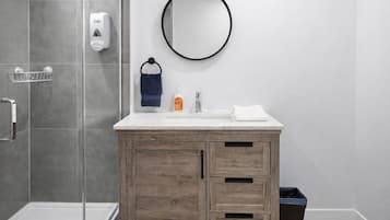 A modern bathroom with a shower, a wooden vanity, a round mirror, and a hand soap dispenser.