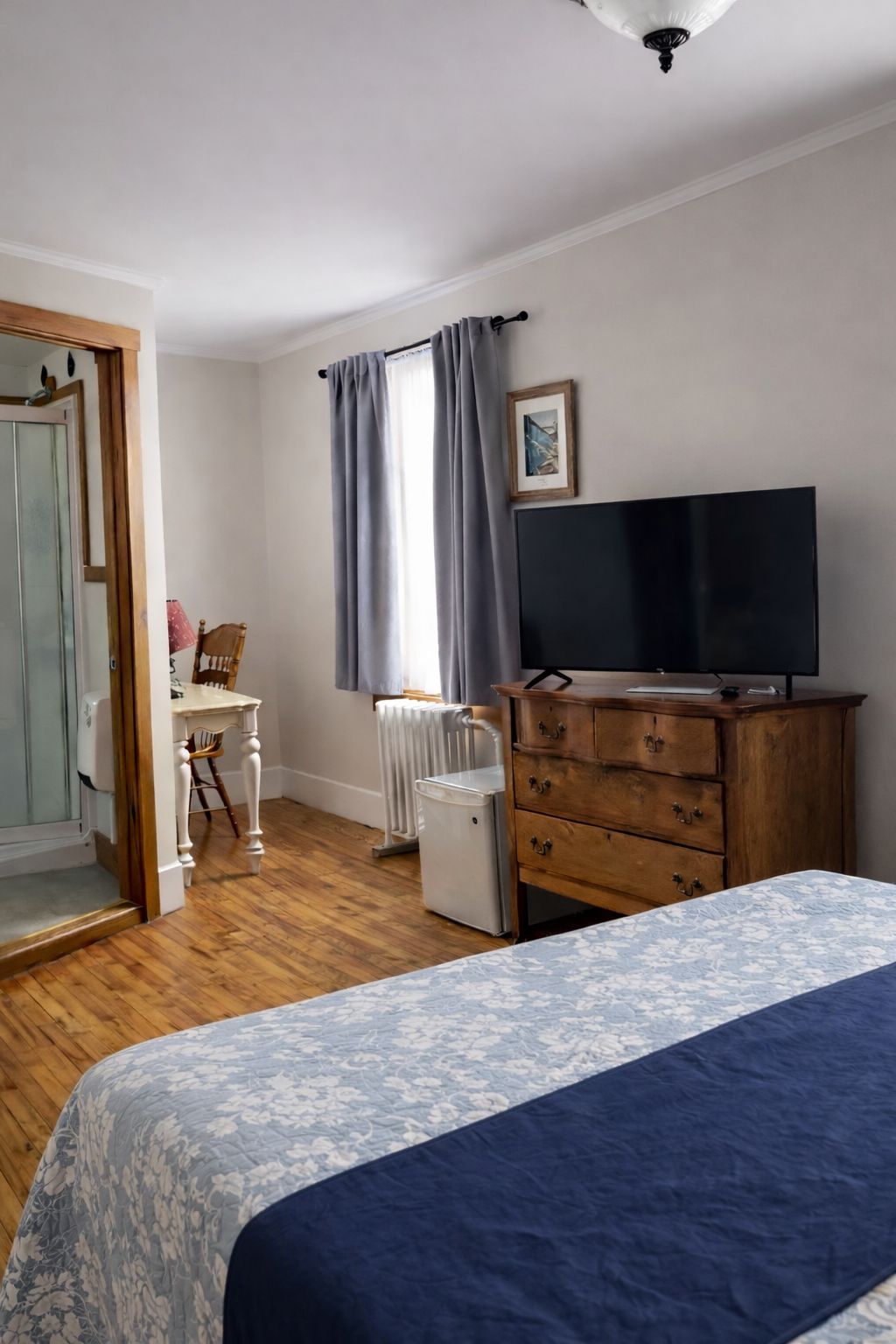 Ett sovrum med en säng, en byrå, en tv, ett element och ett fönster med gardiner.