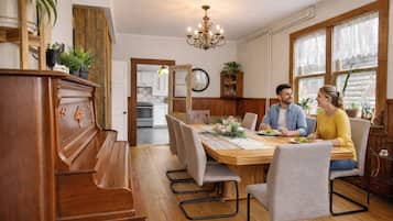 A dining area with a wooden table, chairs, and a chandelier. There is a kitchen in the background.