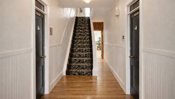A hallway with a wooden floor, white wainscoting, and a staircase with patterned carpet.