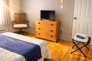 A room with a bed, a wooden dresser with a television on top, a chair, and a small table with towels. - Auberge Orpailleur chambre 302 (Val-d'Or)