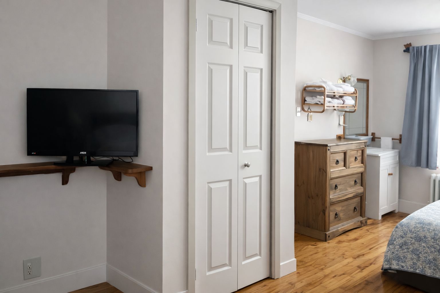 Une chambre à coucher avec un téléviseur posé sur une étagère en bois, une armoire, une commode et une salle de bain équipée d’un miroir et d’un porte-serviettes.