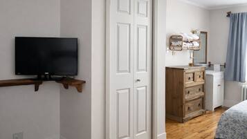 Une chambre à coucher avec un téléviseur posé sur une étagère en bois, une armoire, une commode et une salle de bain équipée d’un miroir et d’un porte-serviettes.