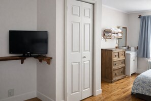 Une chambre à coucher avec un téléviseur posé sur une étagère en bois, une armoire, une commode et une salle de bain équipée d’un miroir et d’un porte-serviettes.